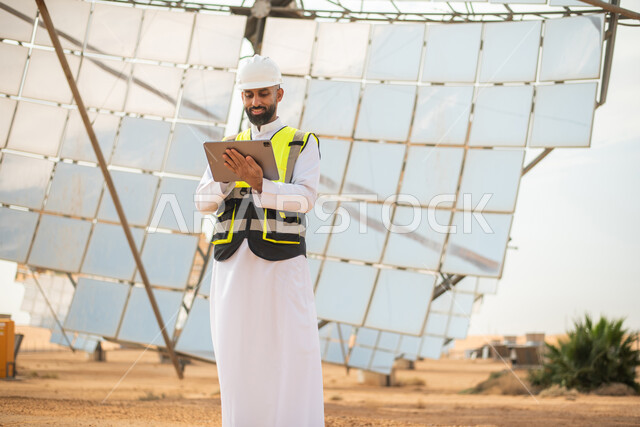 الواح وستلايت انتاج الطاقة الكهربائية , صورة من الاسفل لمهندس عربي خليجي سعودي يشرف على محطات الطاقة الشمسية في السعودية، استخدام جهاز التابلت في ادارة المشاريع , إرتداء خوذة وسترة الحماية الخاصة بالعمل، مخططات وتصاميم هندسية، محطات الطاقة الشمسية المتجدد