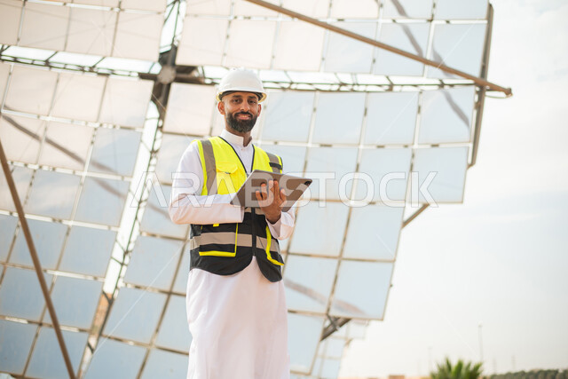 النظر الى الكاميرا , صورة من الاسفل لمهندس عربي خليجي سعودي يشرف على محطات الطاقة الشمسية في السعودية، استخدام جهاز التابلت في ادارة المشاريع , إرتداء خوذة وسترة الحماية الخاصة بالعمل، مخططات وتصاميم هندسية،  محطات الطاقة الشمسية المتجددة