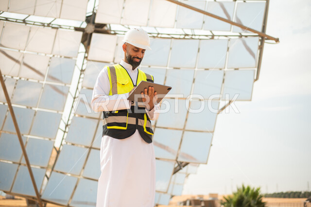 صورة من الاسفل لمهندس عربي خليجي سعودي يشرف على محطات الطاقة الشمسية في السعودية، استخدام جهاز التابلت في ادارة المشاريع , إرتداء خوذة وسترة الحماية الخاصة بالعمل، مخططات وتصاميم هندسية، محطات الطاقة الشمسية المتجددة، الواح وستلايت انتاج الطاقة الكهربائية