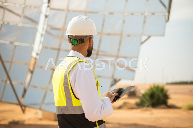صورة مقربة لمهندس عربي خليجي سعودي يشرف على محطات الطاقة الشمسية في السعودية، استخدام جهاز التابلت في ادارة المشاريع , إرتداء خوذة وسترة الحماية الخاصة بالعمل، مخططات وتصاميم هندسية، محطات الطاقة الشمسية المتجددة، الواح وستلايت انتاج الطاقة الكهربائية 