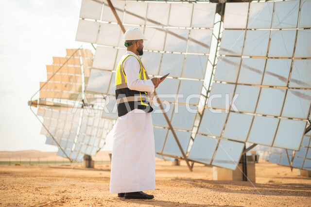 مهندس عربي خليجي سعودي يشرف على محطات الطاقة الشمسية في السعودية، استخدام جهاز التابلت في ادارة المشاريع , إرتداء خوذة وسترة الحماية الخاصة بالعمل، مخططات وتصاميم هندسية، محطات الطاقة الشمسية المتجددة، الواح وستلايت انتاج الطاقة الكهربائية