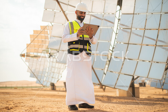  استخدام جهاز التابلت في ادارة المشاريع , صورة من الاسفل لمهندس عربي خليجي سعودي يشرف على محطات الطاقة الشمسية في السعودية، إرتداء خوذة وسترة الحماية الخاصة بالعمل، مخططات وتصاميم هندسية،محطات الطاقة الشمسية المتجددة، الواح وستلايت انتاج الطاقة الكهربائية