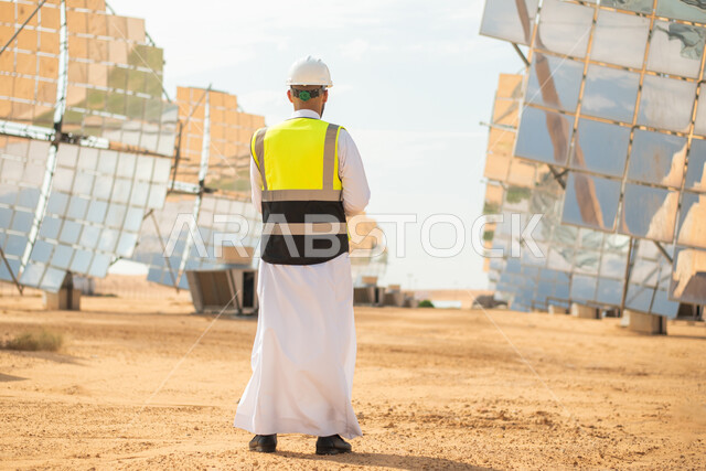 صورة من الخلف لمهندس عربي خليجي سعودي يشرف على محطات الطاقة الشمسية في السعودية، صورة الجسم بالطول الكامل , إرتداء خوذة وسترة الحماية الخاصة بالعمل، مخططات وتصاميم هندسية، محطات الطاقة الشمسية المتجددة، الواح وستلايت انتاج الطاقة الكهربائية