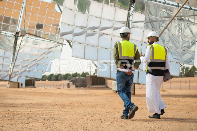 صورة من الخلف لمهندسان عربيان خليجيان سعوديان يشرفان على محطات الطاقة الشمسية في السعودية، إيماءات تدل على المشي , إرتداء خوذة وسترة الحماية الخاصة بالعمل، مخططات وتصاميم هندسية، حوارات ونقاشات عمل ،  محطات الطاقة الشمسية المتجددة، الواح وستلايت انتاج الط