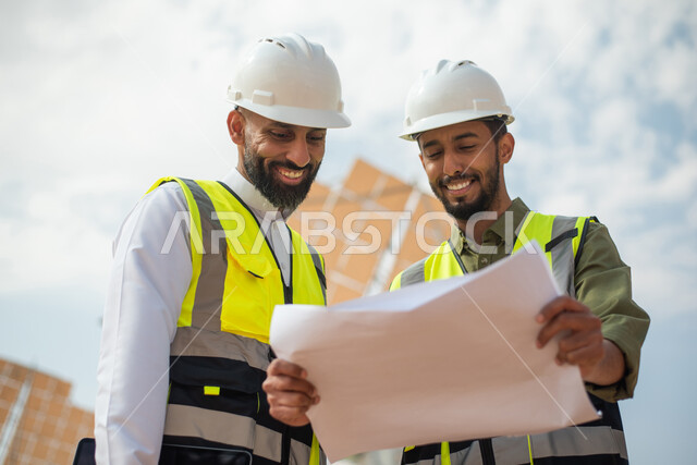 صورة من الاسفل لمهندسان عربيان خليجيان سعوديان يشرفان على محطات الطاقة الشمسية في السعودية، إرتداء خوذة وسترة الحماية الخاصة بالعمل، مخططات وتصاميم هندسية، حوارات ونقاشات عمل ،  محطات الطاقة الشمسية المتجددة، الواح وستلايت انتاج الطاقة الكهربائية , تطور ا