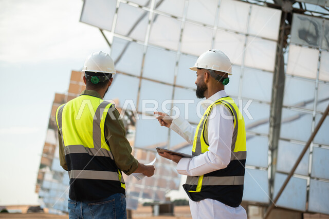 ألواح طاقة , مهندسان عربيان خليجيان سعوديان يشرفان على محطات الطاقة الشمسية ، إرتداء خوذة وسترة الحماية ، الإشراف على العمل، ستلايت، أنظمة الطاقة الشمسية، الطاقة المتجددة. 