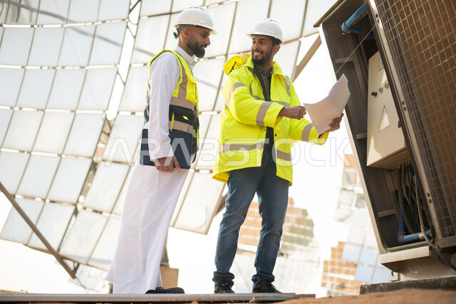 مخططات وتصاميم هندسية، مهندسان عربيان خليجيان سعوديان يشرفان على محطات الطاقة الشمسية في السعودية، إرتداء خوذة وسترة الحماية الخاصة بالعمل، صورة الجسم بالطول الكامل , حوارات ونقاشات عمل ،  محطات الطاقة الشمسية المتجددة، الواح وستلايت انتاج الطاقة الكهربائ