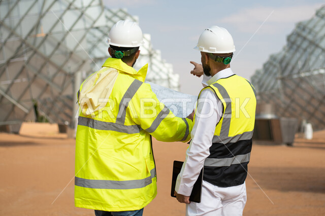 Two Saudi Arabian Gulf energy engineers supervising solar power plants in Saudi Arabia, dialogues and discussions, wearing a helmet and work protection jacket, engineering plans and designs, renewable energy, clean energy, natural energy, satellite, solar