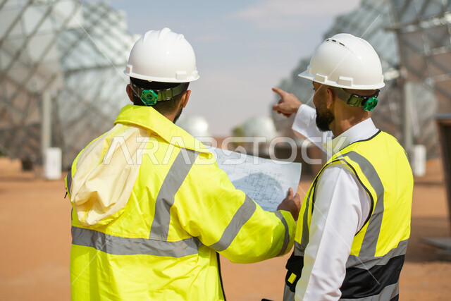 حوارات ونقاشات عمل ، صورة من الخلف لمهندسان عربيان خليجيان سعوديان يشرفان على محطات الطاقة الشمسية في السعودية، إرتداء خوذة وسترة الحماية الخاصة بالعمل، مخططات وتصاميم هندسية، محطات الطاقة الشمسية المتجددة، الواح وستلايت انتاج الطاقة الكهربائية , تطور الت