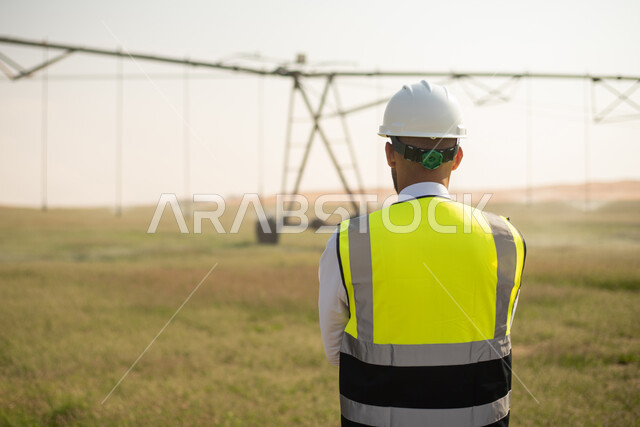 صورة من الخلف لمهندس زراعي عربي خليجي سعودي يقف متكتف اليدين ,النظر الى الكاميرا  ، مفهوم الهندسة الزراعية ,   استخدام التقنية والتكنولوجيا لتطوير مستقبل القطاع الزراعي , تحسين انتاج المحاصيل الزراعية في السعودية , تنميه المزارع والحقول الخضراء .