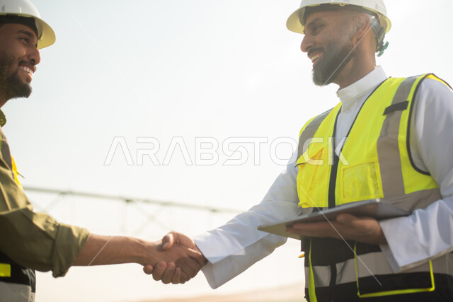 صورة مقربة لمهندسان زراعيان عربيان خليجيان سعوديان في حقل زراعي ، إرتداء خوذة وسترة الحماية، إتفاقيات و صفقات عمل، استخدام التقنية والتكنولوجيا الحديثة في القطاع الزراعي  ، آلات ومعدات الري المستقبلي, خلفية الاراضي الزراعية .