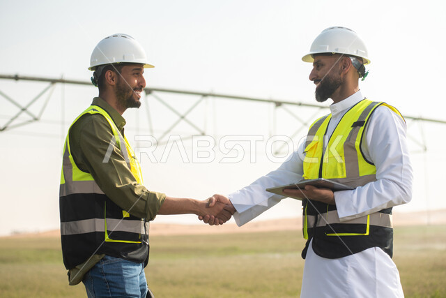 مهندسان زراعيان عربيان خليجيان سعوديان في حقل زراعي ، إرتداء خوذة وسترة الحماية، إتفاقيات و صفقات عمل، استخدام التقنية والتكنولوجيا الحديثة في القطاع الزراعي  ، آلات ومعدات الري المستقبلي, خلفية الاراضي الزراعية .