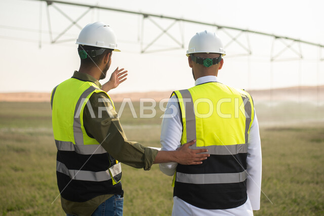  إتفاقيات و صفقات عمل، صورة من الخلف لمهندسان زراعيان عربيان خليجيان سعوديان في حقل زراعي ، إرتداء خوذة وسترة الحماية،  نقاشات واجتماع عمل , استخدام التقنيه والتكنولوجيا في الهندسة الزراعية  ، آلات ومعدات الري المستقبلي.