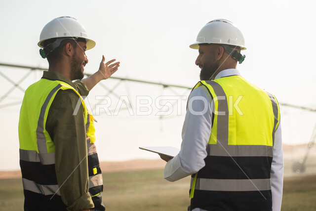 صورة من الخلف لمهندسان زراعيان عربيان خليجيان سعوديان في حقل زراعي ، إرتداء خوذة وسترة الحماية، إتفاقيات و صفقات عمل، نقاشات واجتماع عمل , استخدام التقنيه والتكنولوجيا في الهندسة الزراعية  ، آلات ومعدات الري المستقبلي.