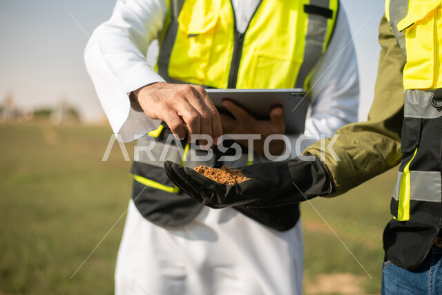 Agricultural lands and fields, two Saudi Arabian Gulf agricultural engineers in an agricultural field, wearing a helmet and protection jacket, business agreements and deals, agricultural lands and fields, soil testing, using a mobile phone (tablet)