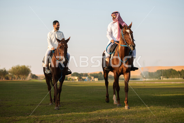 أنشطة ترفيهية مع الحيوانات ، مزرعة تدريب وترويض الخيول في السعودية ، تربية الخيول في الاسطبل ، هواية ركوب الخيل في نادي الفروسية ، رجلان عربيان خليجيان سعوديان يرتديان الزي السعودي التقليدي يمتطيان الخيل العربي الأصيل ، أنشطة ترفيهية وقضاء أوقات ممتعة