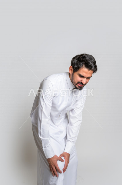 بورتريه لرجل عربي سعودي خليجي يرتدي الثوب السعودي, إيماءات وجه ويدين تدل على الشعور بالوجع , ألم بالقدم , الإحساس بالتعب والإرهاق , الحاجه للعلاج , طلب المساعده , خلفية بيضاء .