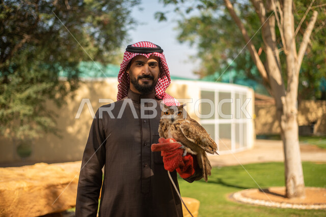  رجل عربي خليجي سعودي يحمل بيده طائر البومة، قضاء أوقات ممتعة في حديقة الحيوانات في المملكة العربية السعودية، أماكن سياحية ترفيهية، أنشطة ترفيهية مع الطيور، تربية وتدريب الطيور الجارحة في المحمية الطبيعية، طائر البومة
