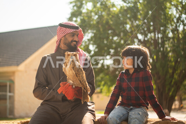 رجل عربي سعودي خليجي  مع إبنه، قضاء أوقات ممتعة في حديقة الحيوانات في المملكة العربية السعودية، أماكن سياحية ترفيهية، أنشطة ترفيهية مع الطيور، تربية وتدريب الطيور الجارحة في المحمية الطبيعية، طائر البومة