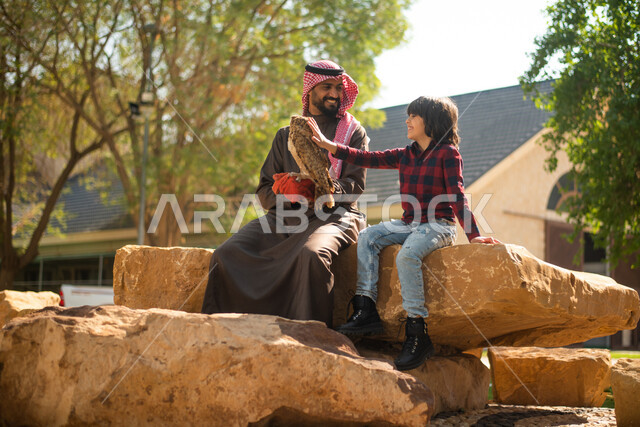 رجل عربي سعودي خليجي  مع إبنه، قضاء أوقات ممتعة في حديقة الحيوانات في المملكة العربية السعودية، أماكن سياحية ترفيهية، أنشطة ترفيهية مع الطيور، تربية وتدريب الطيور الجارحة في المحمية الطبيعية، طائر البومة