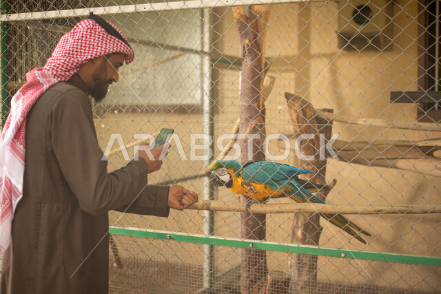 رجل عربي خليجي سعودي يرتدي الزي التقليدي, التقاط صور تذكارية عبر الهاتف المحمول, قضاء أوقات ممتعة في حديقة الحيوانات في المملكة العربية السعودية، أماكن سياحية ترفيهية بالسعودية، محمية طبيعية، طائر الببغاء، فعاليات ترفيهية عائلية