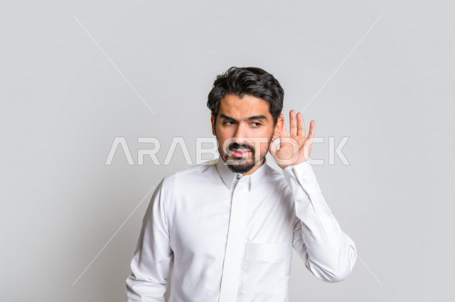 بورتريه لرجل عربي سعودي خليجي يرتدي الثوب السعودي, إيماءات يد تدل على الإنصات, حسن الإستماع, التركيز على الكلام, محاولة التجسس والتنصت, سماع الأسرار , خلفية بيضاء .