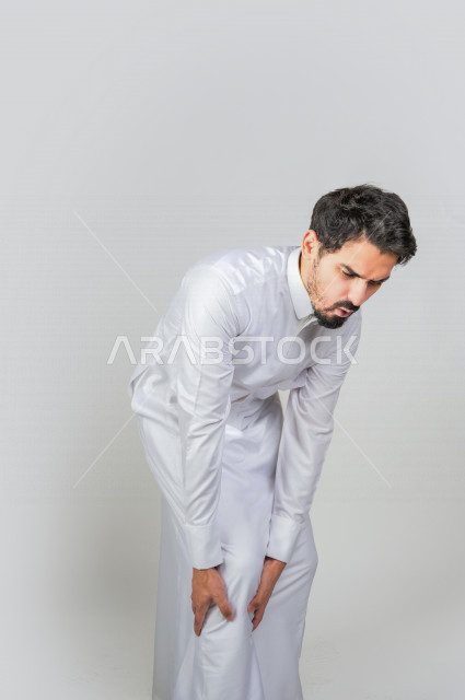 بورتريه لرجل عربي سعودي خليجي يرتدي الثوب السعودي, إيماءات وجه ويدين تدل على الشعور بالوجع , ألم بالقدم , الإحساس بالتعب والإرهاق , الحاجه للعلاج , طلب المساعده , خلفية بيضاء .