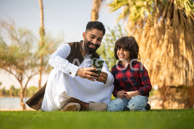 Using a mobile phone, a Saudi Arabian Gulf young man and child, looking at the mobile phone, enjoying playing, spending fun times in a public park in the Kingdom of Saudi Arabia, recreational tourist places in Saudi Arabia, family entertainment activities