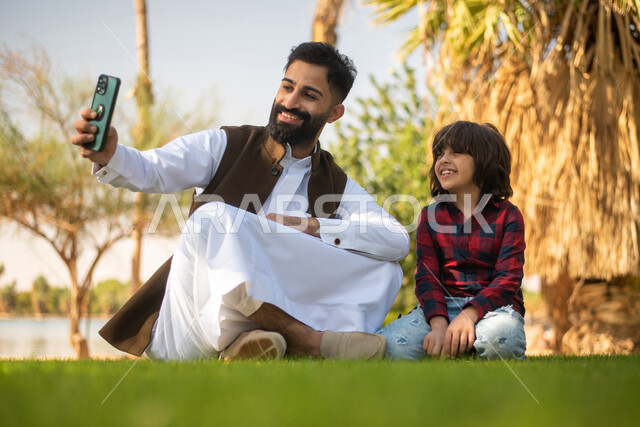 طفل ووالده عربيان سعوديان خليجيان , التقاط صور سيلفي عبر الهاتف المحمول, الاستمتاع باللعب , قضاء أوقات ممتعة في احد الحدائق العامة في المملكة العربية السعودية، أماكن سياحية ترفيهية بالسعودية، فعاليات ترفيهية عائلية