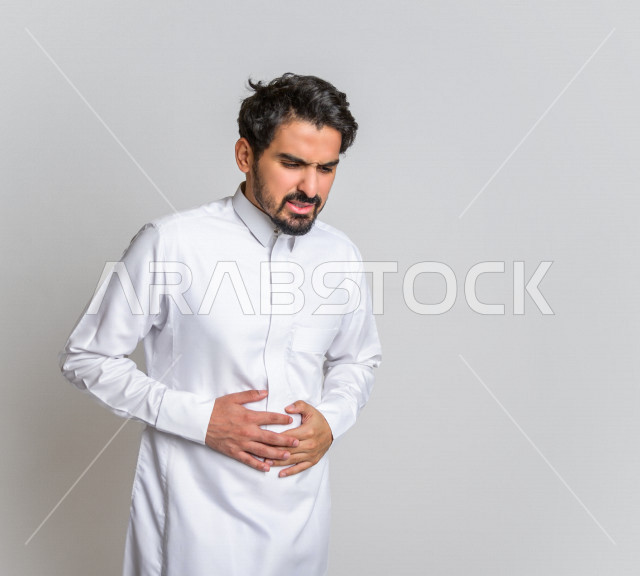 بورتريه لرجل عربي سعودي خليجي يرتدي الثوب السعودي, إيماءات وجه ويدين تدل على الشعور بالتعب , ألم البطن , الإحساس بالمرض والتسمم , التعب والإرهاق , أوجاع القولون , الحاجه للعلاج , طلب المساعده , خلفية بيضاء .