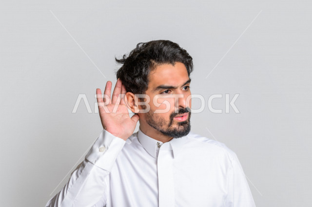 بورتريه لرجل عربي سعودي خليجي يرتدي الثوب السعودي, إيماءات يد تدل على الإنصات, حسن الإستماع, التركيز على الكلام, محاولة التجسس والتنصت, سماع الأسرار , خلفية بيضاء .
