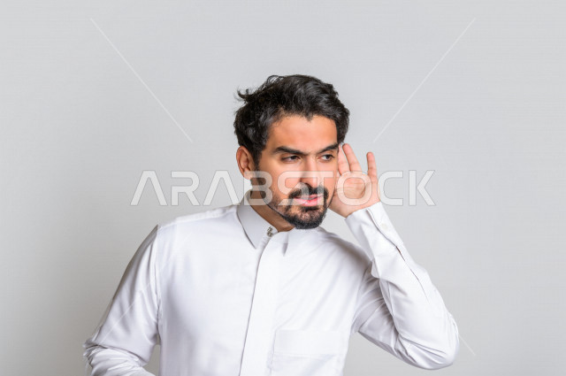 بورتريه لرجل عربي سعودي خليجي يرتدي الثوب السعودي, إيماءات يد تدل على الإنصات, حسن الإستماع, التركيز على الكلام, محاولة التجسس والتنصت, سماع الأسرار , خلفية بيضاء .