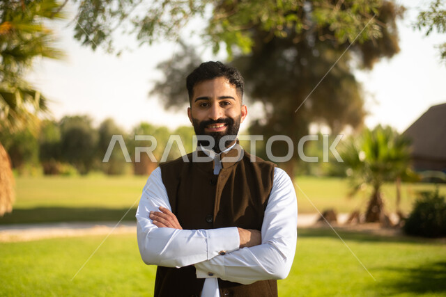رجل عربي خليجي سعودي يقف متكتف اليدين في إحدى الحدائق العامة في المملكة العربية السعودية, ينظر الى الكاميرا،  يقف باستقامه ،قضاء وقت ممتع في الحدائق, ايماءات تدل على السعادة والفرح , مزارع واستراحات .