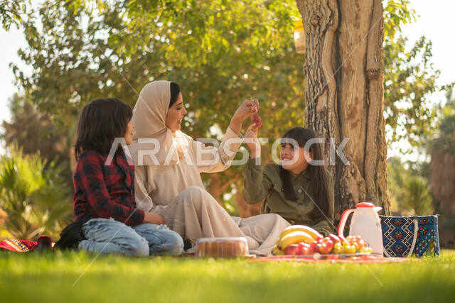 A Saudi Arabian Gulf woman spends pleasant times with her children, spending pleasant times in one of the public parks in Saudi Arabia, taking a picnic in the open air, sitting in the green nature.