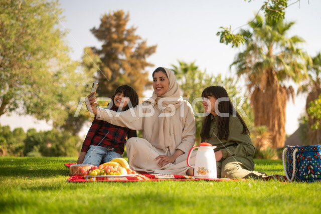 أجواء عائلية سعيدة قضاء أوقات ممتعة في إحدى الحدائق بالسعودية ، مرأة عربية خليجية سعودية برفقة أبناءها ، نزهة في في الهواء الطلق ، الجلوس في الطبيعة الخضراء ، عطلة صيفية.