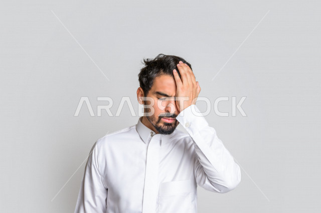 بورتريه لرجل عربي سعودي خليجي يرتدي الثوب السعودي, إيماءات وجه ويدين تدل على الشعور بالصداع, ألم بالرأس, الإحساس بالمرض, التعب والإرهاق, الحاجه للعلاج, آلام الشقيقة والصداع النصفي , خلفية بيضاء .