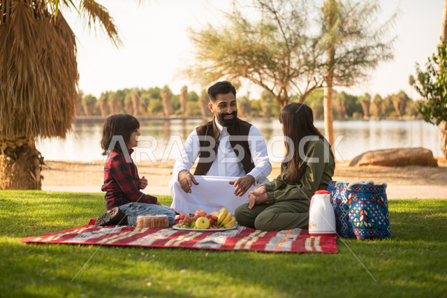 A Saudi Arabian Gulf man accompanied by his children, spending pleasant times in one of the public parks in Saudi Arabia, picnicking in the open air, sitting in the green nature, summer vacation.