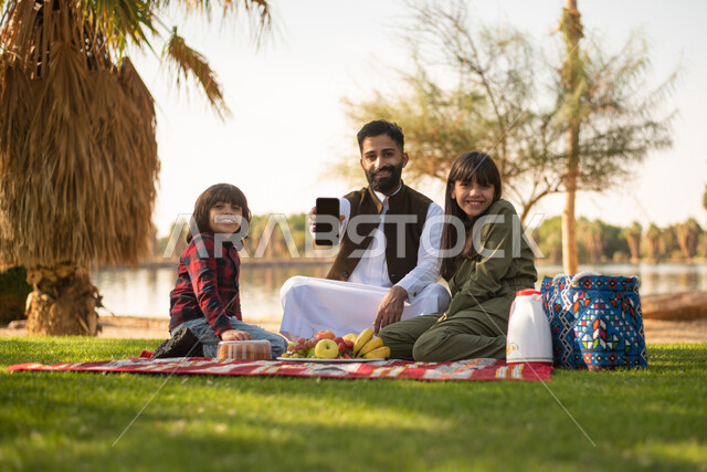 رجل  سعودي عربي خليجي برفقة أبناءه ،  شاشة الهاتف المحمول السوداء ،  قضاء اوقات ممتعة في إحدى الحدائق العامة بالسعودية ، نزهة في في الهواء الطلق ، الجلوس في الطبيعة الخضراء ، عطلة صيفية.