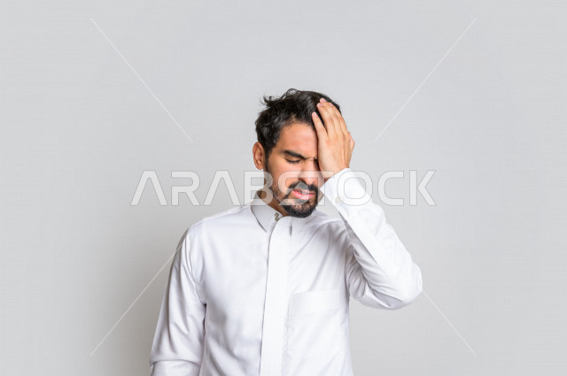 بورتريه لرجل عربي سعودي خليجي يرتدي الثوب السعودي, إيماءات وجه ويدين تدل على الشعور بالصداع, ألم بالرأس, الإحساس بالمرض, التعب والإرهاق, الحاجه للعلاج, آلام الشقيقة والصداع النصفي , خلفية بيضاء .