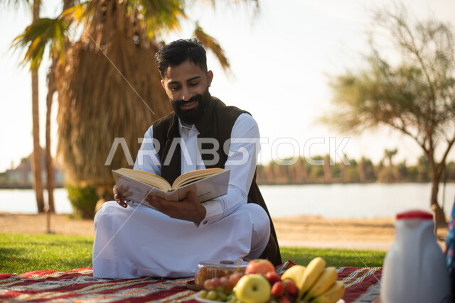 رجل عربي خليجي سعودي يستمتع بقرأة القصص والروايات في إحدى الحدائق العامة بالسعودية ، نزهة في الهواء الطلق ، عطلة صيفية ، الجلوس في الطبيعة الخضراء.