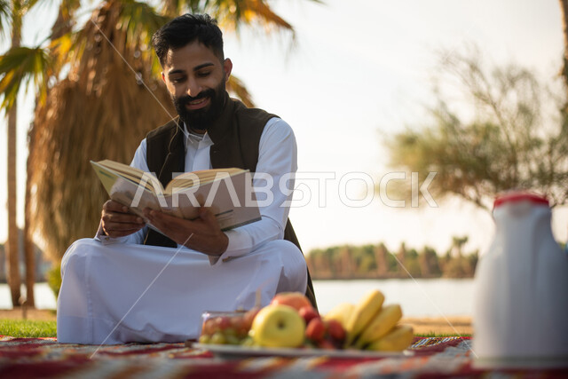 رجل سعودي عربي خليجي يستمتع بقرأة القصص والروايات في إحدى الحدائق العامة بالسعودية ، نزهة في الهواء الطلق ، عطلة صيفية ، الجلوس في الطبيعة الخضراء.