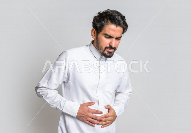 بورتريه لرجل عربي سعودي خليجي يرتدي الثوب السعودي, إيماءات وجه ويدين تدل على الشعور بالتعب , ألم البطن , الإحساس بالمرض والتسمم , التعب والإرهاق , أوجاع القولون , الحاجه للعلاج , طلب المساعده , خلفية بيضاء .