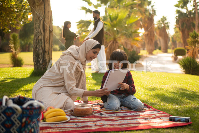 نزهة في الهواء الطلق ، امرأة سعودية عربية خليجية برفقة ولدها  في إحدى الحدائق العامة بالسعودية ، ممارسة هواية الرسم والكتابة ، عطلة صيفية ، الجلوس في الطبيعة الخضراء.