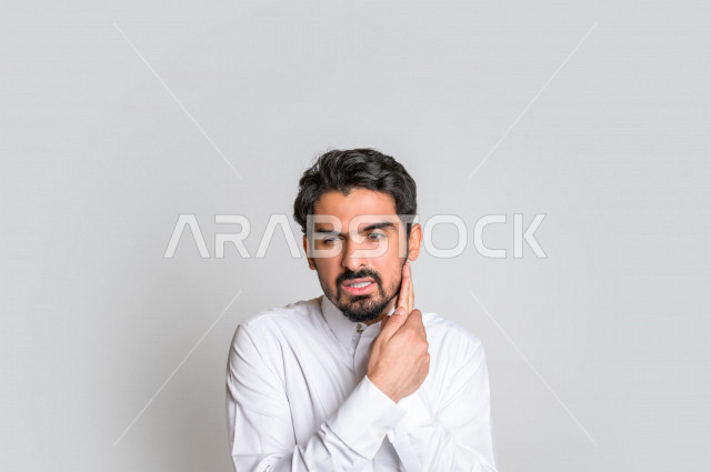 بورتريه لرجل عربي سعودي خليجي باللباس السعودي التقليدي , يقوم بإيماءات وجه و يدين تدل على الألم والوجع , تسوس الأضراس, التعب والإرهاق, طلب العلاج و المساعده, الحاجه لزيارة طبيب الأسنان , ألم الأسنان , الإنتفاخ والإلتهاب بالفم واللثة , خلفية بيضاء