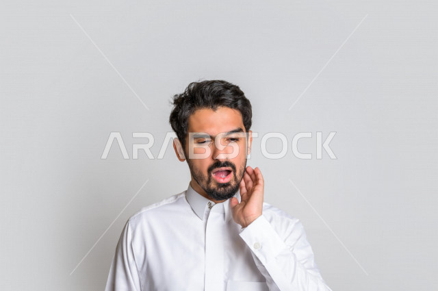 Portrait of a Saudi Arabian Gulf man in traditional Saudi dress, making facial and hands gestures indicating pain and pain, molar decay, fatigue and exhaustion, seeking treatment and help, the need to visit a dentist, toothache, bloating and inflammation