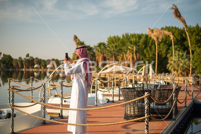 A picture of a Saudi Arab Gulf man taking souvenir photos using a mobile phone, a picnic in the open air, spending enjoyable times in a tourist place, a water lake, recreational outings in the Kingdom of Saudi Arabia.