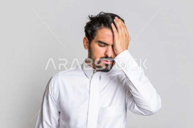 بورتريه لرجل عربي سعودي خليجي يرتدي الثوب السعودي, إيماءات وجه ويدين تدل على الشعور بالصداع, ألم بالرأس, الإحساس بالمرض, التعب والإرهاق, الحاجه للعلاج, آلام الشقيقة والصداع النصفي , خلفية بيضاء .