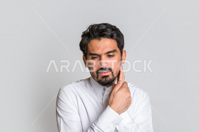 بورتريه لرجل عربي سعودي خليجي باللباس السعودي التقليدي , يقوم بإيماءات وجه و يدين تدل على الألم والوجع , تسوس الأضراس, التعب والإرهاق, طلب العلاج و المساعده, الحاجه لزيارة طبيب الأسنان , ألم الأسنان , الإنتفاخ والإلتهاب بالفم واللثة , خلفية بيضاء