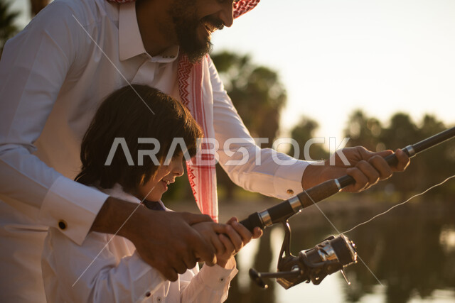 تصوير مقرب لرجل سعودي  خليجي  عربي يقوم بتعليم ولده هواية الصيد ، ممارسة الأنشطة الترفيهية في الهواء الطلق ،  رحلة صيد الأسماك ، عطلة صيفية ، حدائق ونزهات ترفيهية في المملكة العربية السعودية.