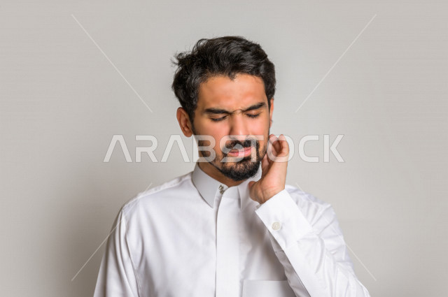 Portrait of a Saudi Arabian Gulf man in traditional Saudi dress, making facial and hands gestures indicating pain and pain, molar decay, fatigue and exhaustion, seeking treatment and help, the need to visit a dentist, toothache, bloating and inflammation